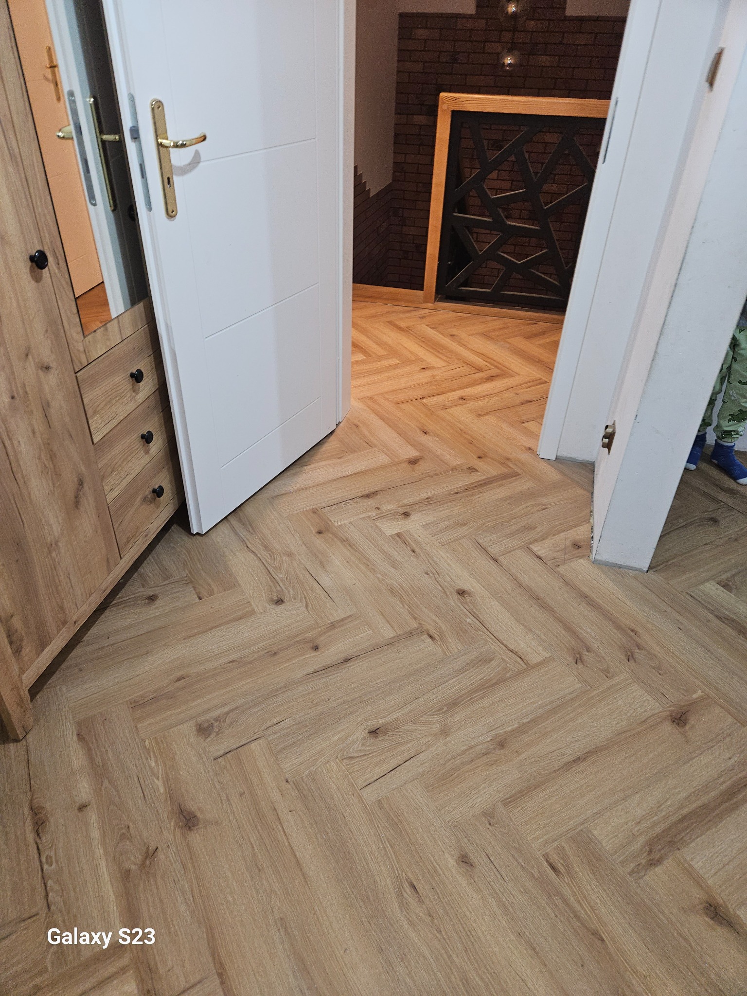 Ułożona podłoga z paneli w jodełkę, widok z perspektywy podłogi, z fragmentem szafy i otwartymi drzwiami do pokoju z ceglaną ścianą w tle.