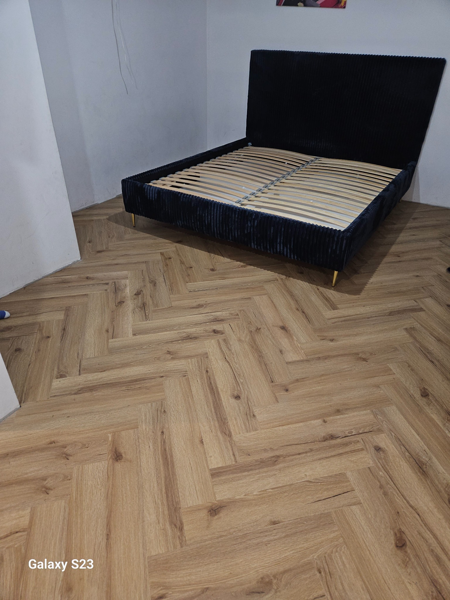 Ułożona podłoga z paneli w jodełkę, na której stoi czarne łóżko z drewnianym stelażem. Widok na fragment ściany i podłogę.