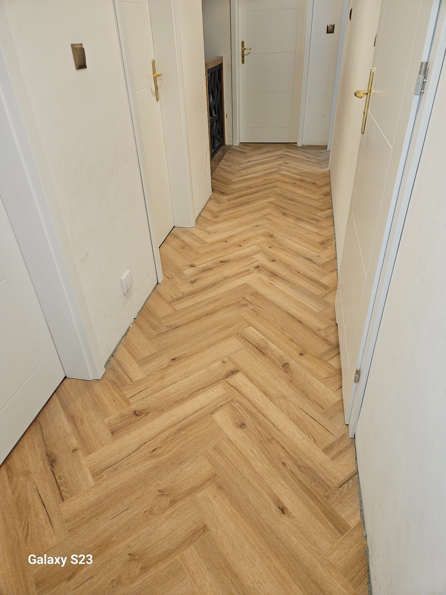 Ułożona podłoga z paneli w jodełkę w korytarzu z białymi drzwiami. Widoczne detale łączeń paneli i wykończenia przy ścianach. Jasne, naturalne oświetlenie.