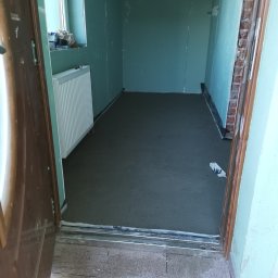 Estetyczny - Wnętrze pomieszczenia w trakcie prac remontowych: świeżo wylana, szara wylewka na podłodze, ściany przygotowane pod malowanie, widoczny fragment grzejnika i otwarte drzwi wejściowe.