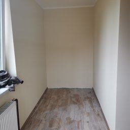 Estetyczny - Wnętrze pokoju po remoncie, widoczne świeżo położone płytki podłogowe imitujące drewno, pomalowane ściany na jasny beż i białe listwy przypodłogowe, grzejnik pod oknem.