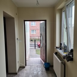 Estetyczny - Wnętrze przedpokoju w trakcie remontu: świeżo pomalowane ściany w odcieniach beżu, ułożona podłoga z płytek imitujących drewno, otwarte drzwi wejściowe, widoczne ślady po pracach na podłodze...