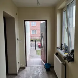 Wnętrze przedpokoju w trakcie remontu: świeżo pomalowane ściany w odcieniach beżu, ułożona podłoga z płytek imitujących drewno, otwarte drzwi wejściowe, widoczne ślady po pracach na podłodze...