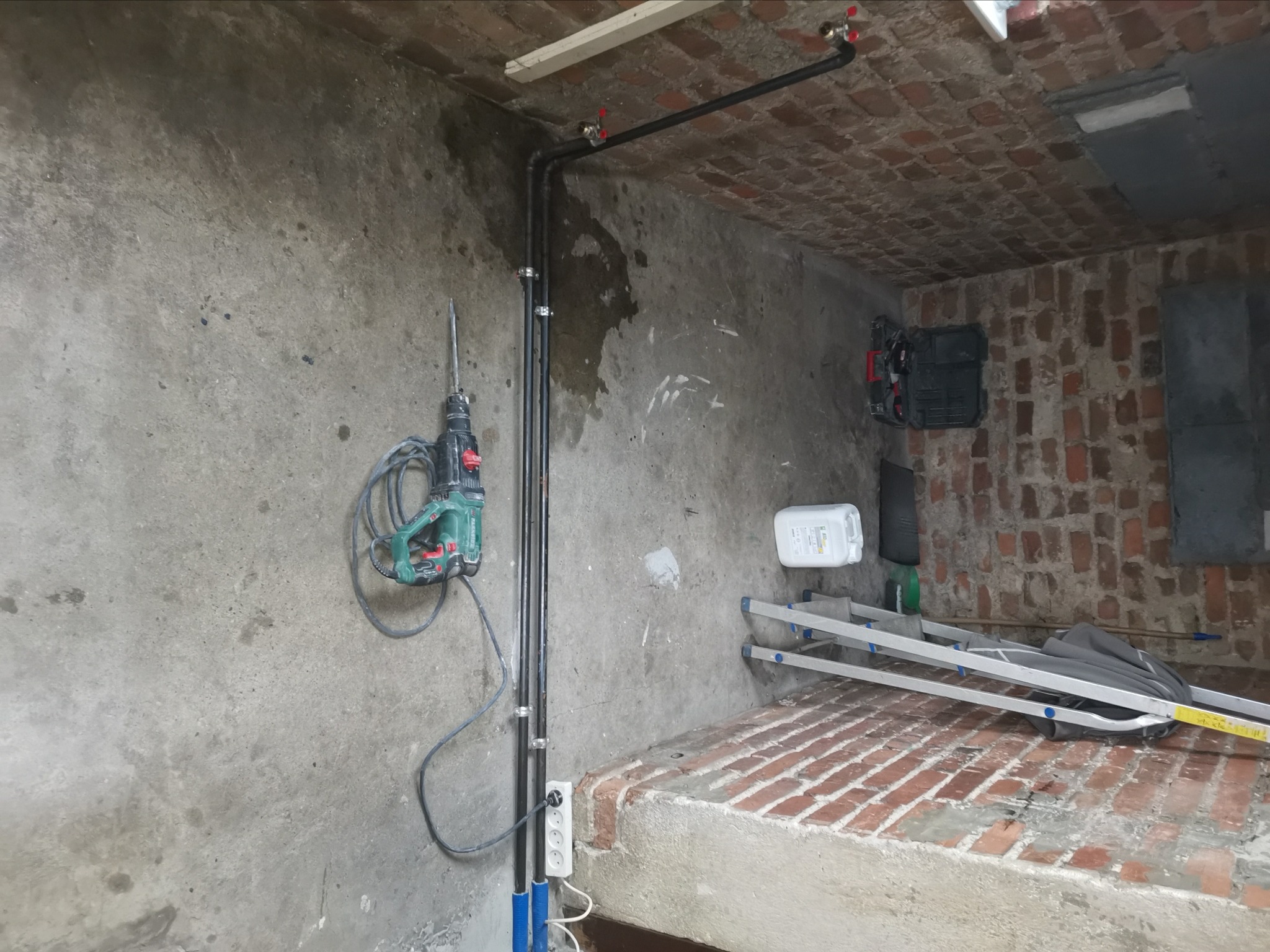Wnętrze piwnicy z surową ścianą, ceglanym podwyższeniem podłogi, rurami biegnącymi wzdłuż ściany, wiszącą wiertarką udarową oraz rozłożoną drabiną opartą o ścianę z cegły.