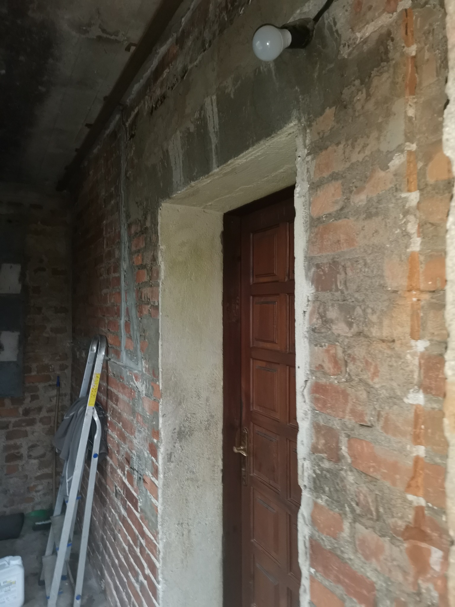 Surowy mur z czerwonej cegły i częściowo otynkowane ściany z widoczną drabiną, ukazujące etap przygotowania do wykończenia wnętrza, z wiszącą żarówką.