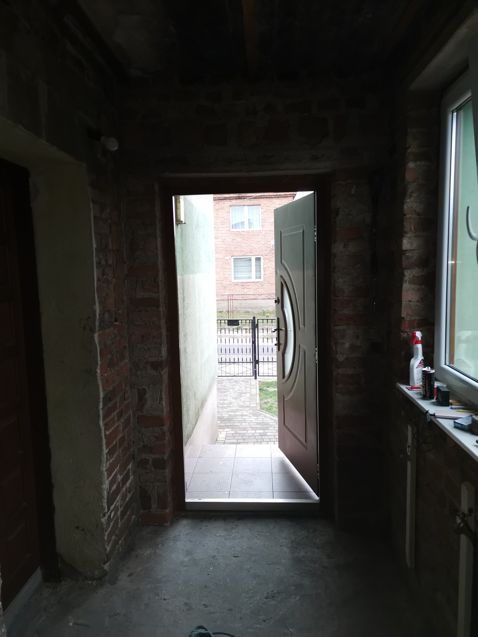 Wnętrze korytarza w trakcie remontu z odsłoniętą cegłą, otwartymi drzwiami wejściowymi i widokiem na podwórze, na parapecie okna widoczne narzędzia i środki czystości.