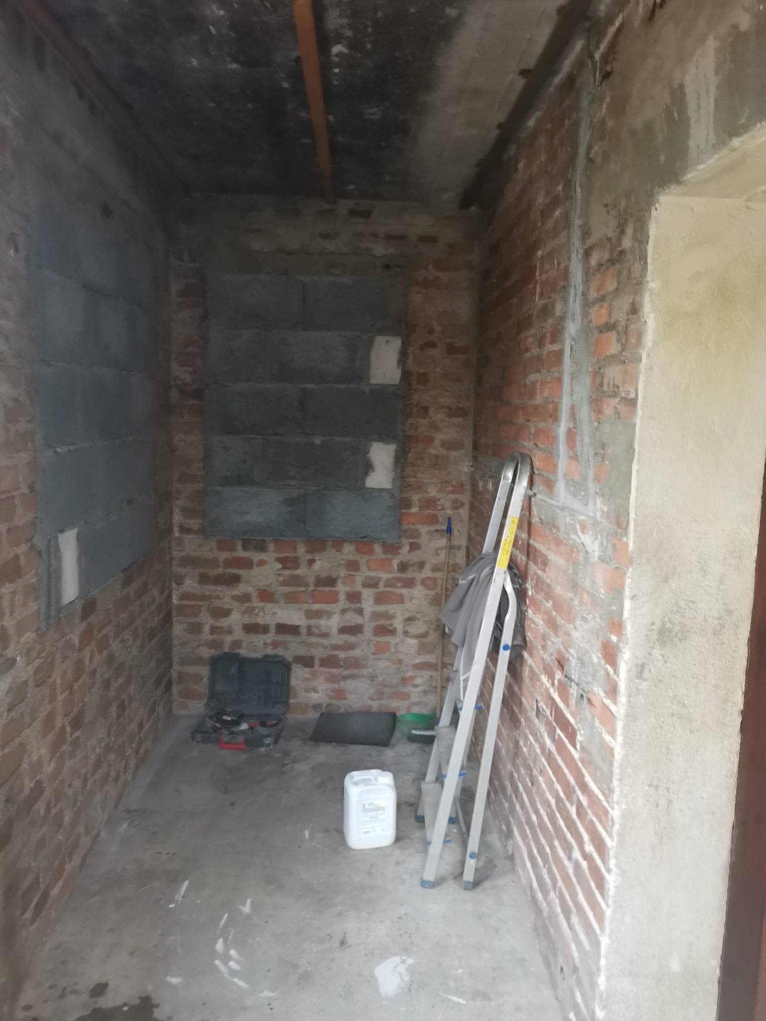 Wnętrze w trakcie remontu: surowe ceglane ściany częściowo pokryte szarymi blokami, aluminiowa drabina oparta o ścianę, narzędzia w czarnym kufrze na podłodze, biały pojemnik z farbą lub gruntem.
