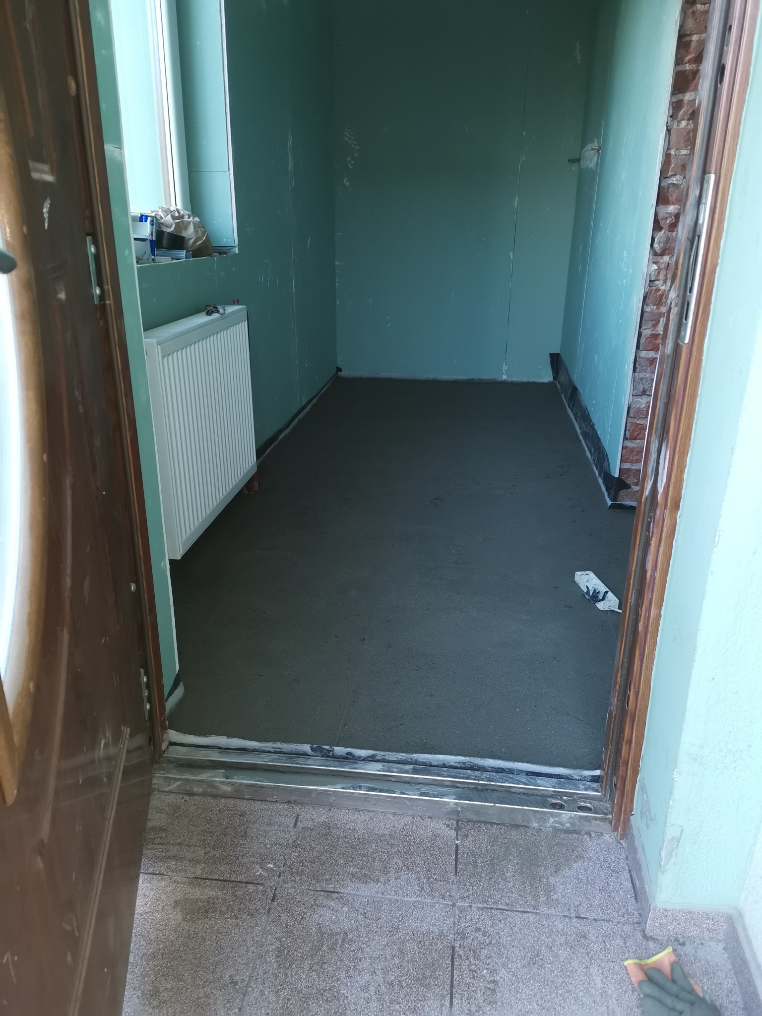 Wnętrze pomieszczenia w trakcie prac remontowych: świeżo wylana, szara wylewka na podłodze, ściany przygotowane pod malowanie, widoczny fragment grzejnika i otwarte drzwi wejściowe.