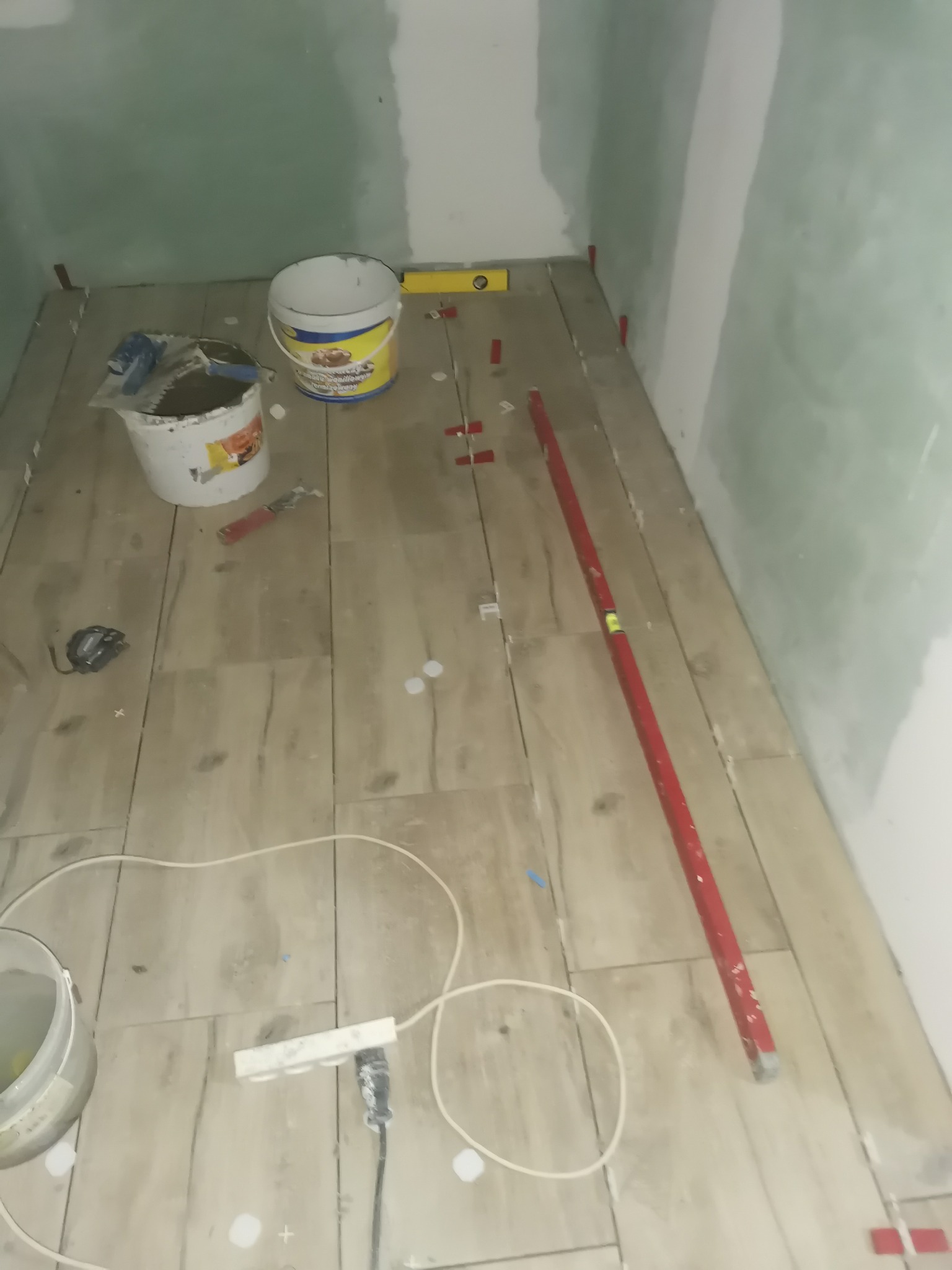 Układanie płytek podłogowych imitujących drewno w rogu pomieszczenia, widoczne narzędzia, poziomica, wiadro z klejem oraz krzyżyki dystansowe.