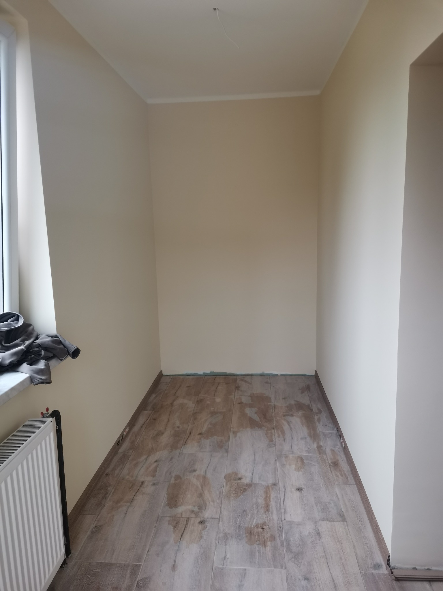 Wnętrze pokoju po remoncie, widoczne świeżo położone płytki podłogowe imitujące drewno, pomalowane ściany na jasny beż i białe listwy przypodłogowe, grzejnik pod oknem.