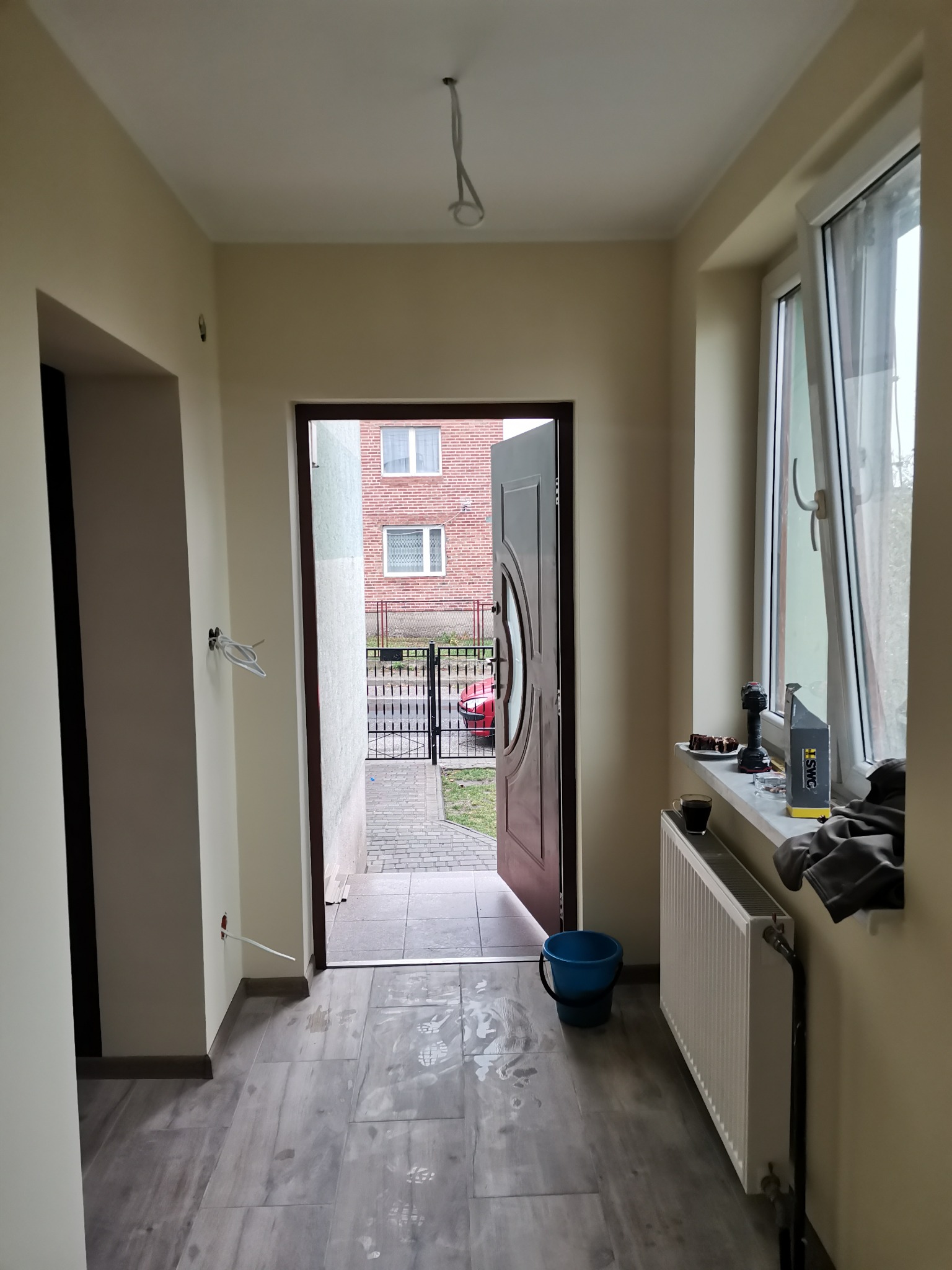 Wnętrze przedpokoju w trakcie remontu: świeżo pomalowane ściany w odcieniach beżu, ułożona podłoga z płytek imitujących drewno, otwarte drzwi wejściowe, widoczne ślady po pracach na podłodze...