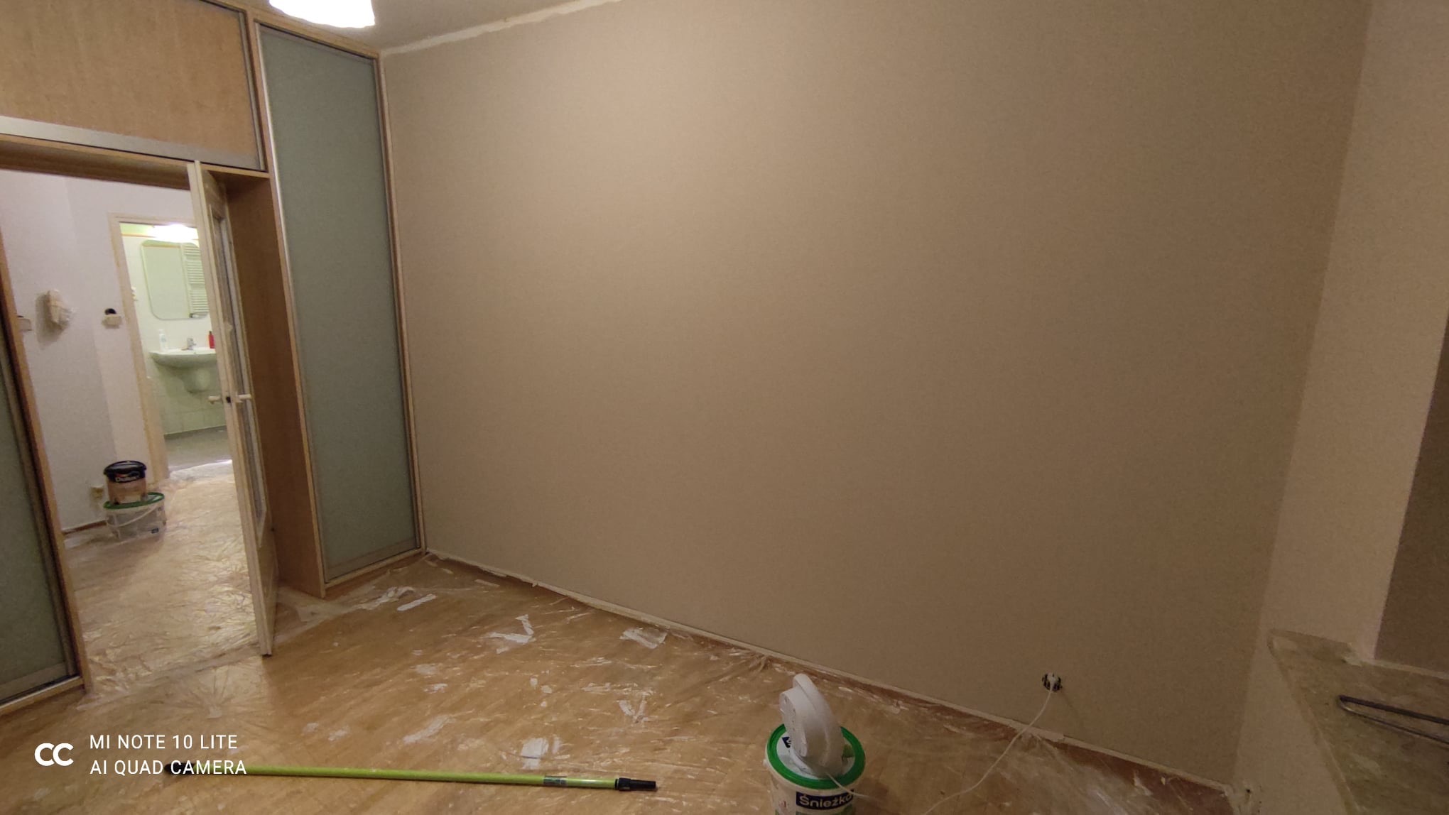 Świeżo pomalowany pokój z beżowymi ścianami, zabezpieczona folią podłoga, widoczny otwarty pokój z umywalką w tle, pędzel i wiadro z farbą Śnieżka na podłodze.