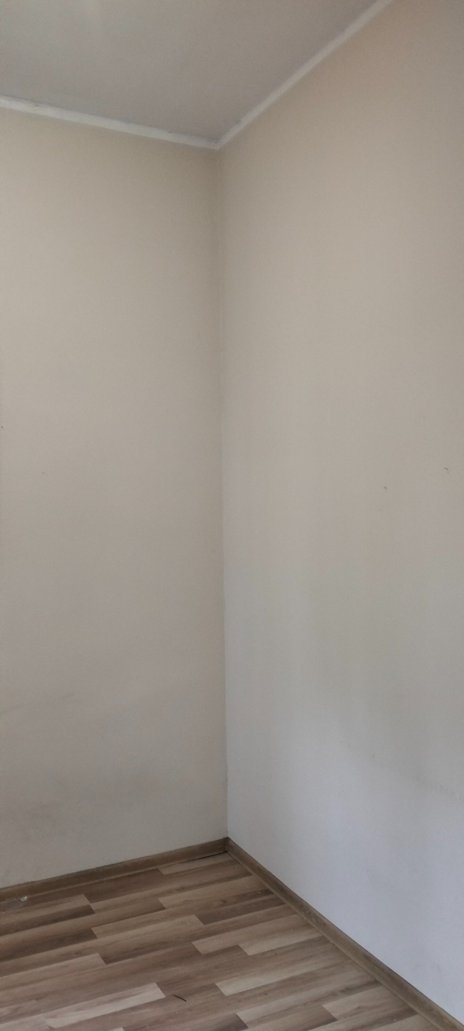 Pusty narożnik pokoju z beżowymi ścianami, białym wykończeniem sufitu i panelami podłogowymi imitującymi drewno. Widoczne ślady zabrudzeń na ścianie przy podłodze.