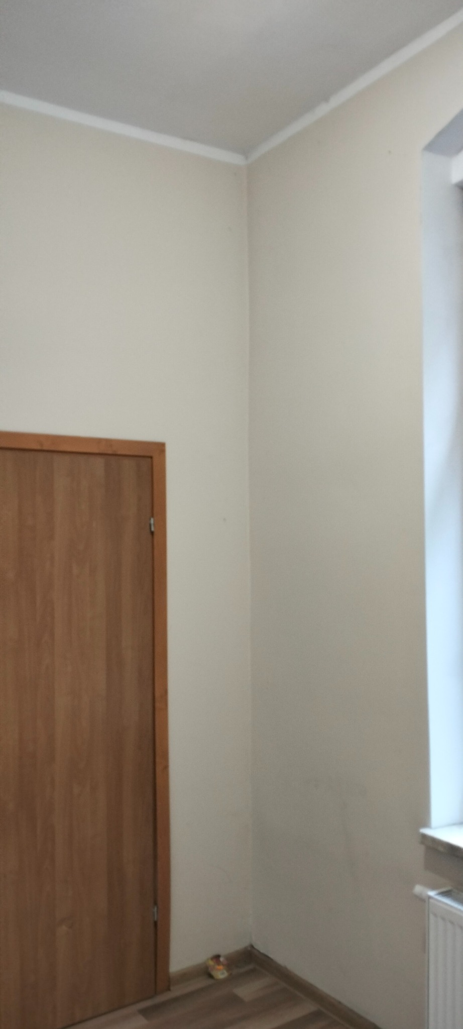 Pomalowane na kremowo ściany w rogu pokoju z drewnianą ramą drzwi po lewej i oknem po prawej stronie, listwa przysufitowa w kolorze białym.
