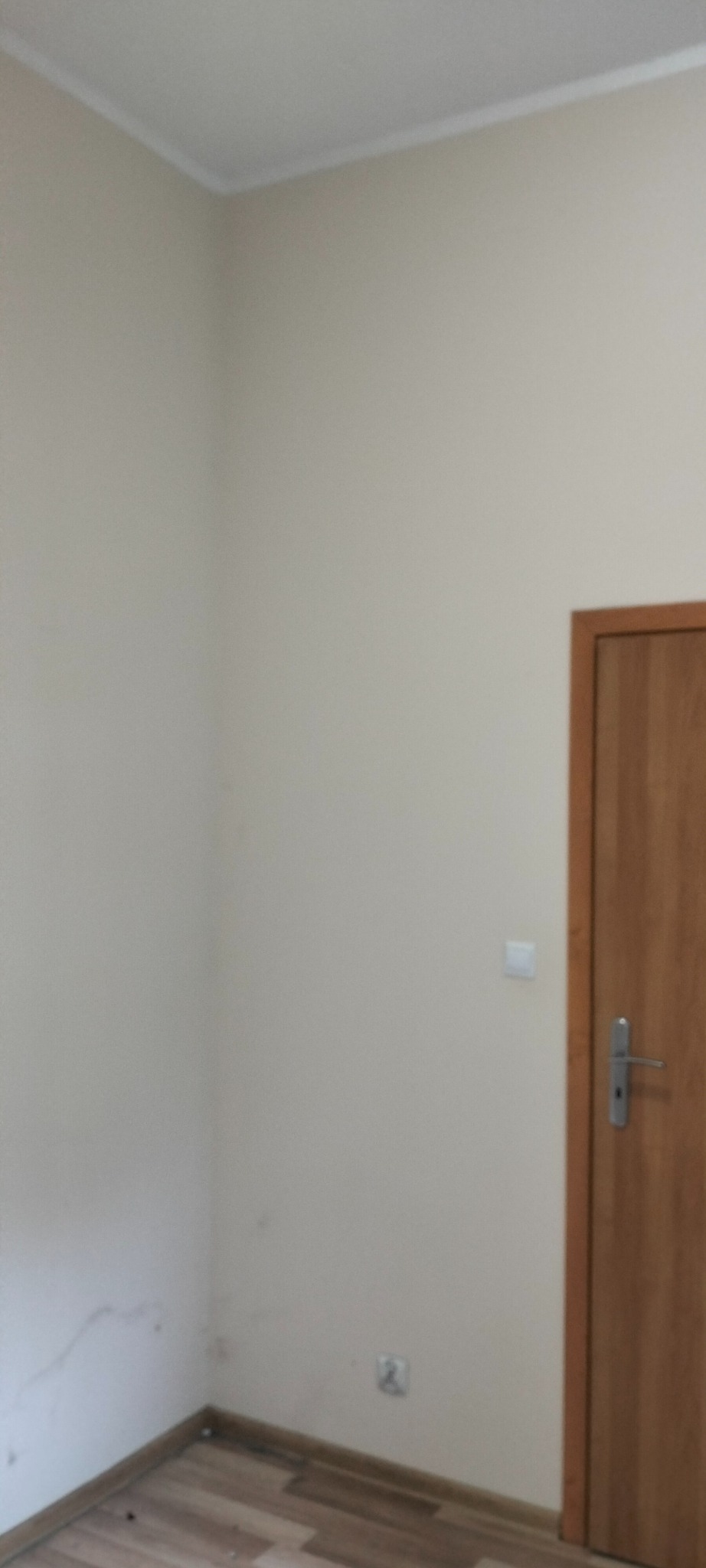 Pomalowane na kremowo ściany pokoju z drewnianą ościeżnicą drzwiową, widoczne gniazdko elektryczne i delikatne zabrudzenia przy podłodze