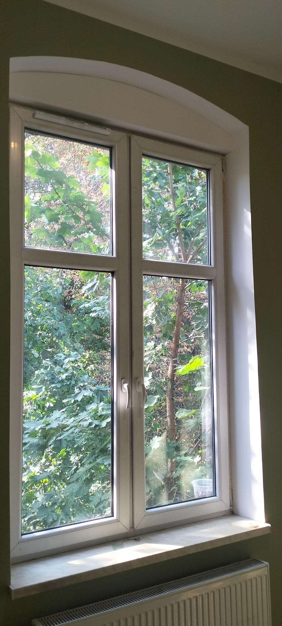 Białe okno PCV z widokiem na zieleń drzew, zamontowane w pokoju z grzejnikiem pod parapetem.