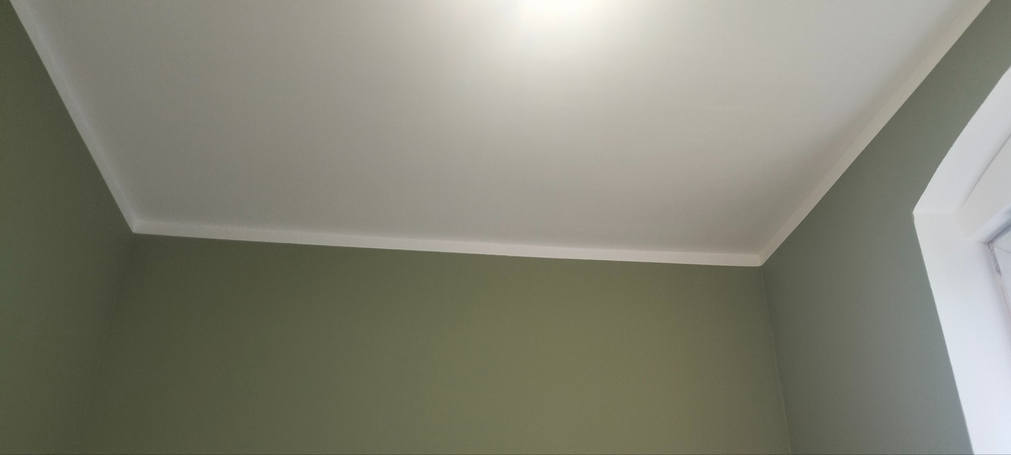 Świeżo pomalowane wnętrze z oliwkowymi ścianami i białym sufitem z listwą przypodłogową, widok z dołu na róg pokoju i ramę okienną.