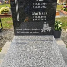 Czarny, polerowany pomnik z wygrawerowanym krzyżem i inskrypcjami, otoczony cmentarnym krajobrazem z kwiatami w donicach.