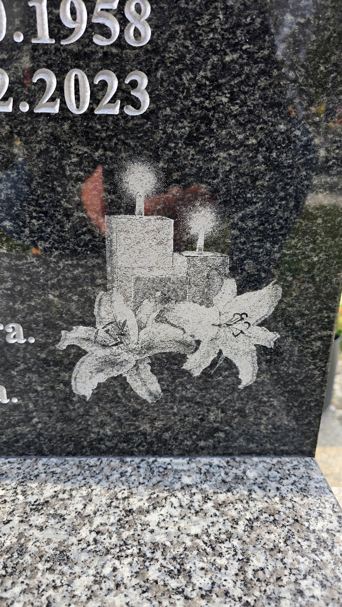 Granitowy pomnik z wygrawerowanymi liliami i świecami, daty 1958-2023 w górnej części.