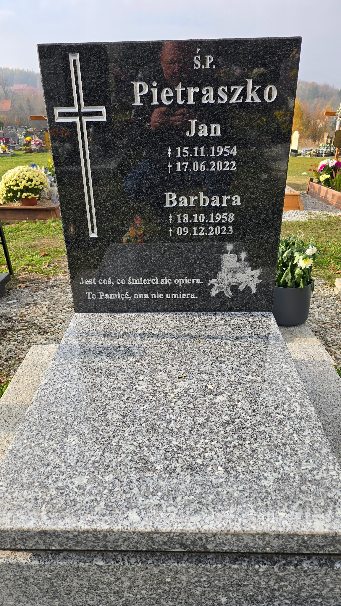 Czarny, polerowany pomnik z wygrawerowanym krzyżem i inskrypcjami, otoczony cmentarnym krajobrazem z kwiatami w donicach.
