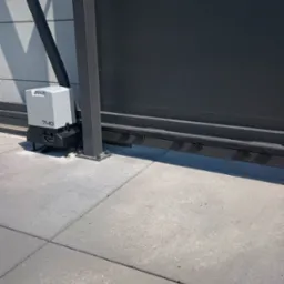 Szary automat bramowy z napędem zębatym, model 740, zamontowany przy nowoczesnej, poziomej bramie przesuwnej na betonowej nawierzchni.
