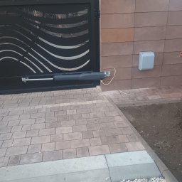 Instel instalacje elektryczne i teletechniczne Tomasz Kozłowicz - Czarna brama wjazdowa z automatyką, widoczny siłownik i sterownik na brązowym murze. Kostka brukowa i kamień ozdobny na podjeździe.