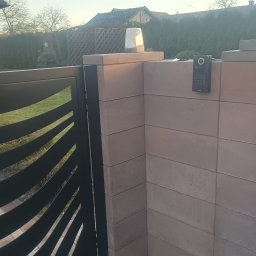 Instel instalacje elektryczne i teletechniczne Tomasz Kozłowicz - Nowoczesna brama z dekoracyjnymi wycięciami, domofon z klawiaturą na murze z cegły, oświetlenie bramy. Widok na ogród i dom w tle.