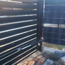 Instel instalacje elektryczne i teletechniczne Tomasz Kozłowicz - Nowoczesna, czarna brama przesuwna z automatyką, widoczny mechanizm napędowy. Częściowo ułożona kostka brukowa w tle siatka ogrodzeniowa.