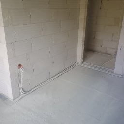 Instel - Ułożone przewody elektryczne w peszlach wzdłuż ścian z białych bloczków i na szarej posadzce, z widocznym wyprowadzeniem z puszki instalacyjnej w rogu pomieszczenia.