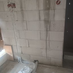 Instel - Instalacja elektryczna w trakcie montażu na ścianie z bloczków, z widocznymi białymi peszlami, puszkami i przewodami. Na ścianie widoczne są odręczne oznaczenia.
