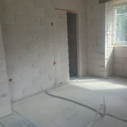 Instel - Wnętrze pomieszczenia w stanie surowym z widoczną instalacją elektryczną: białe przewody biegnące po ścianach z bloczków i betonowej posadzce, zakończone pomarańczowymi puszkami. Naturalne światło...