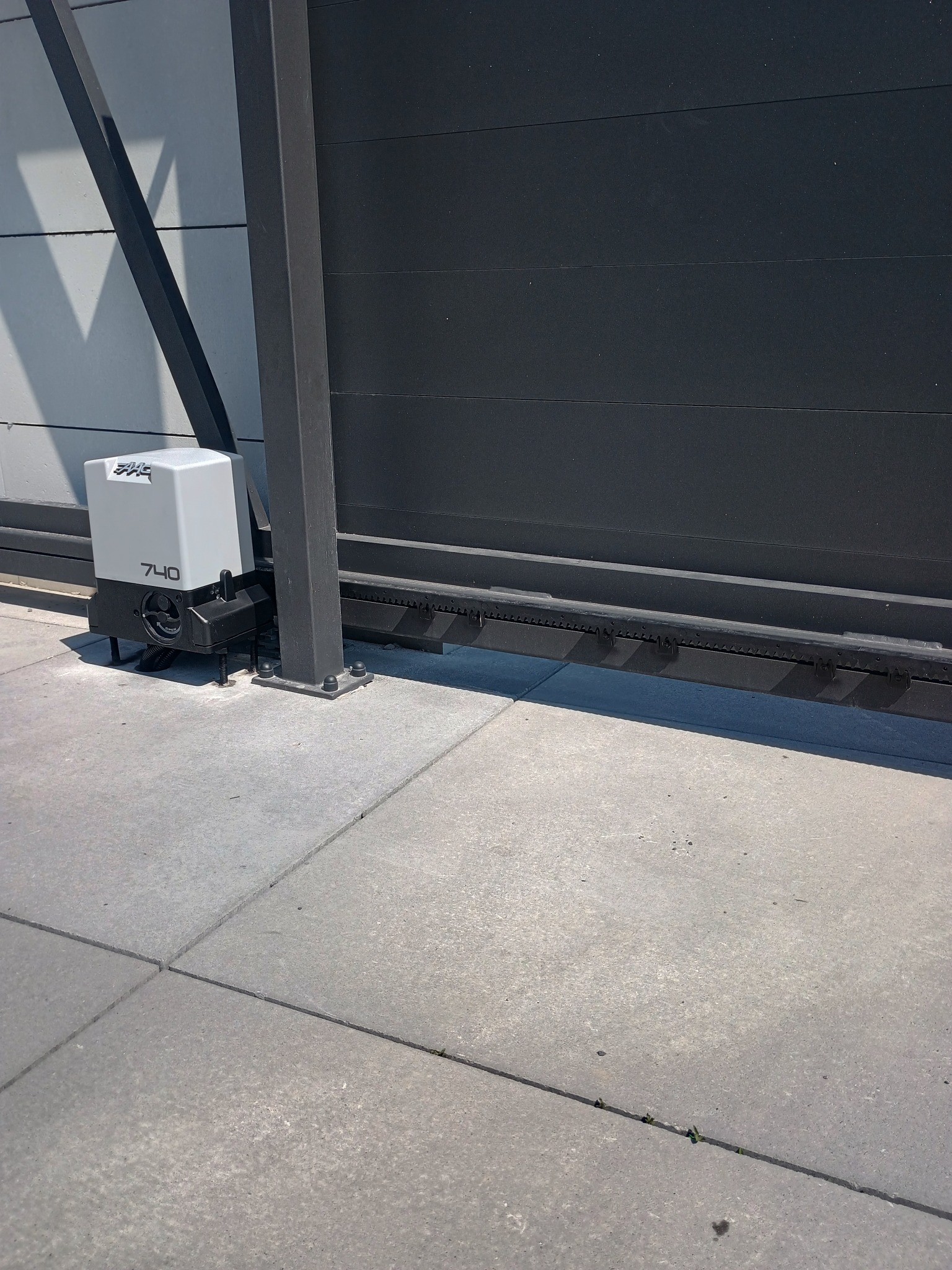 Szary automat bramowy z napędem zębatym, model 740, zamontowany przy nowoczesnej, poziomej bramie przesuwnej na betonowej nawierzchni.