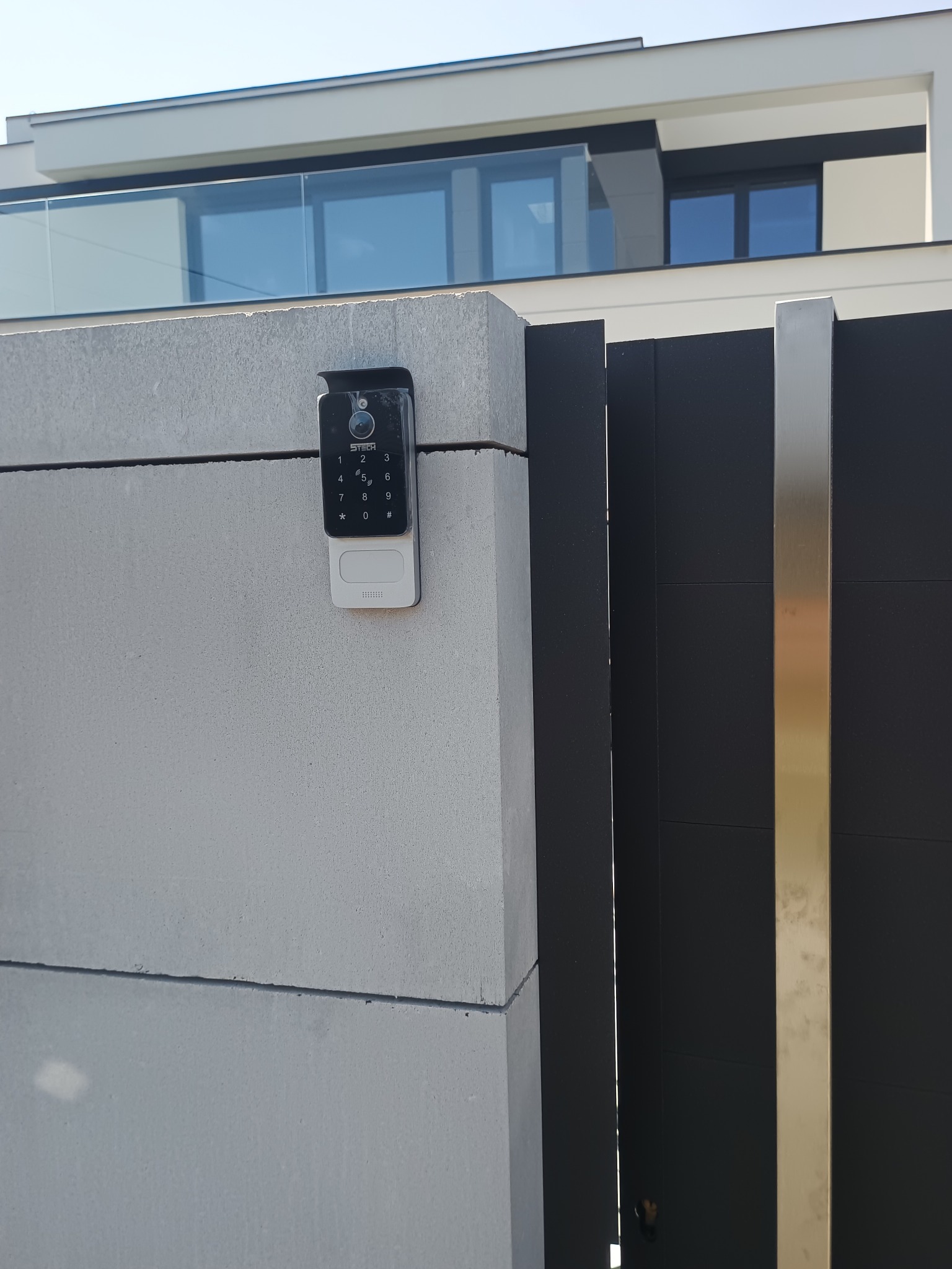 Czarny wideodomofon z klawiaturą numeryczną zamontowany na betonowym słupku ogrodzenia, obok czarnej bramy z elementami ze stali nierdzewnej. Nowoczesny design.