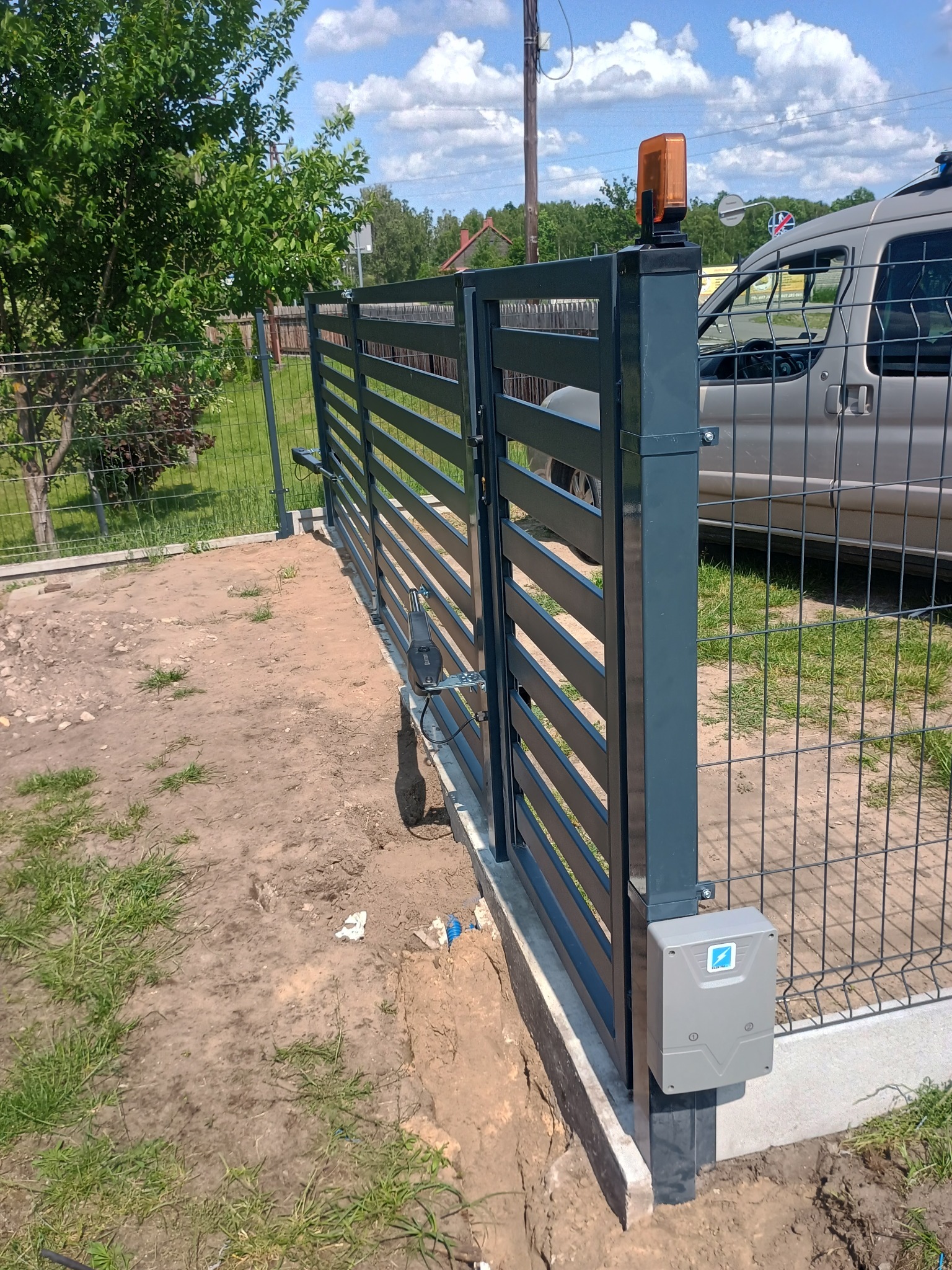 Szara, pozioma brama automatyczna z zamontowanym napędem i lampą ostrzegawczą. Widoczne ogrodzenie panelowe i samochód w tle.