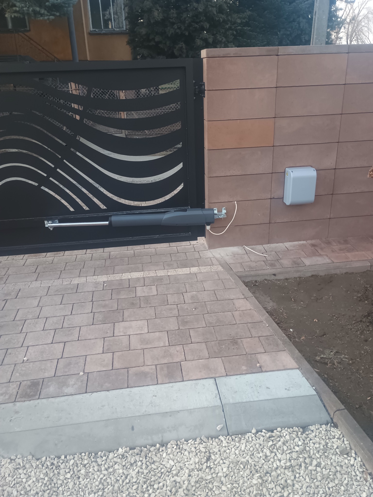 Czarna brama wjazdowa z automatyką, widoczny siłownik i sterownik na brązowym murze. Kostka brukowa i kamień ozdobny na podjeździe.