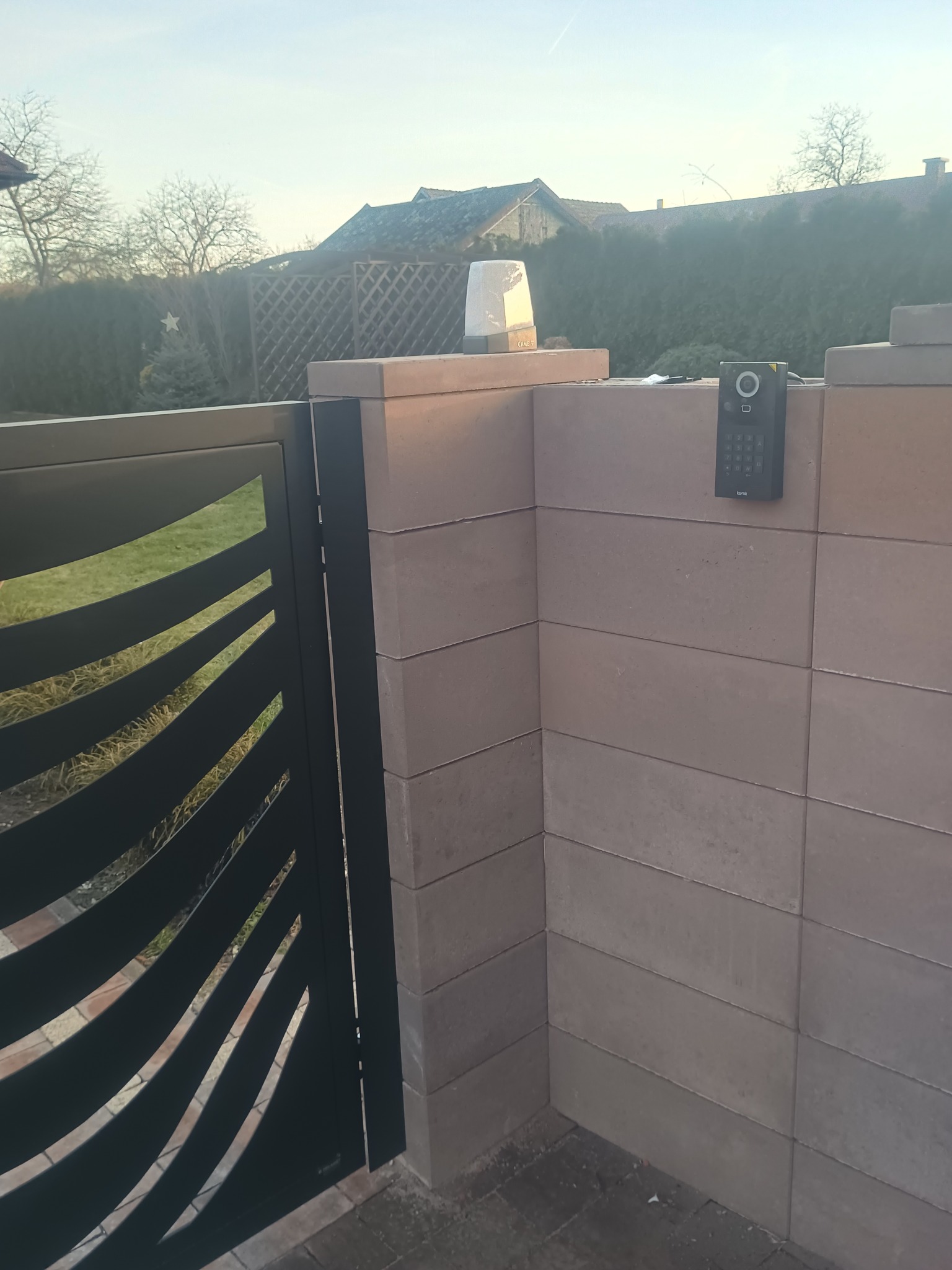 Nowoczesna brama z dekoracyjnymi wycięciami, domofon z klawiaturą na murze z cegły, oświetlenie bramy. Widok na ogród i dom w tle.