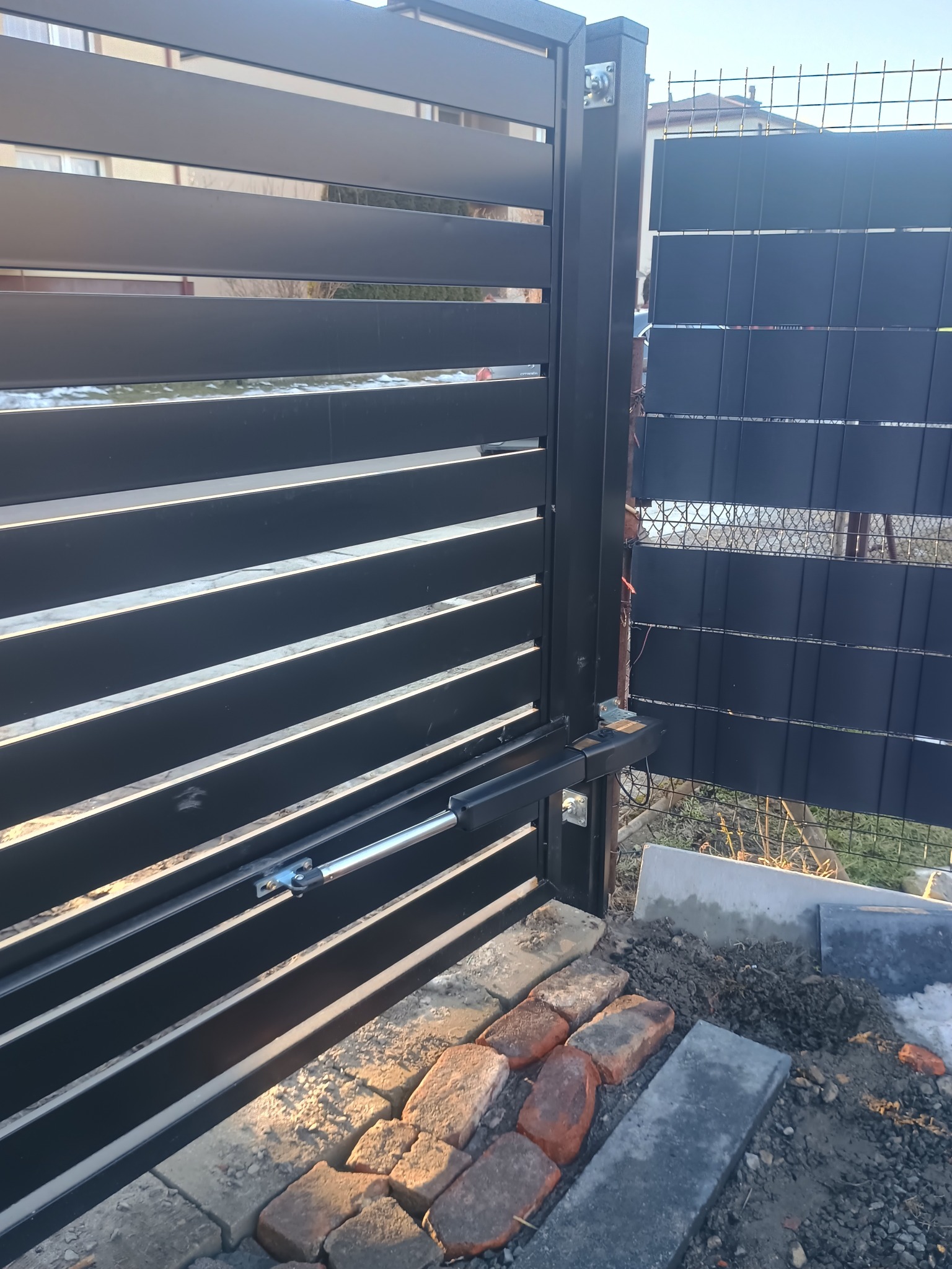 Nowoczesna, czarna brama przesuwna z automatyką, widoczny mechanizm napędowy. Częściowo ułożona kostka brukowa w tle siatka ogrodzeniowa.