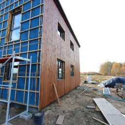 Wood Escape Homes - Dwuetapowa budowa domu szkieletowego: ocieplenie z membraną i drewniana elewacja. Widoczne rusztowanie i teren budowy. Ujęcie z dołu, perspektywa zbieżna.