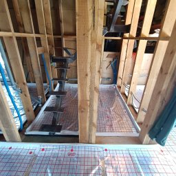 Wood Escape Homes - Konstrukcja drewniana poddasza z instalacją ogrzewania podłogowego na folii refleksyjnej z czerwoną siatką, widoczne rury instalacyjne i fragment schodów.