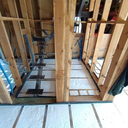 Wood Escape Homes - Wnętrze domu w trakcie budowy szkieletowej: drewniana konstrukcja, izolacja podłogi, instalacje wodne. Widoczne schody metalowe i elementy konstrukcyjne dachu.
