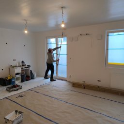 Wood Escape Homes - Malowanie sufitu w jasnym pokoju z zabezpieczoną podłogą. Widoczny malarz z wałkiem na teleskopie, narzędzia i osłonięte okna. Etap prac wykończeniowych.