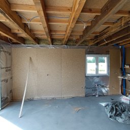 Wood Escape Homes - Wnętrze domu w trakcie budowy: ściany z płyty OSB, izolacja, drewniany strop z instalacjami. Widoczne okno i narzędzia. Surowy stan wykończenia.