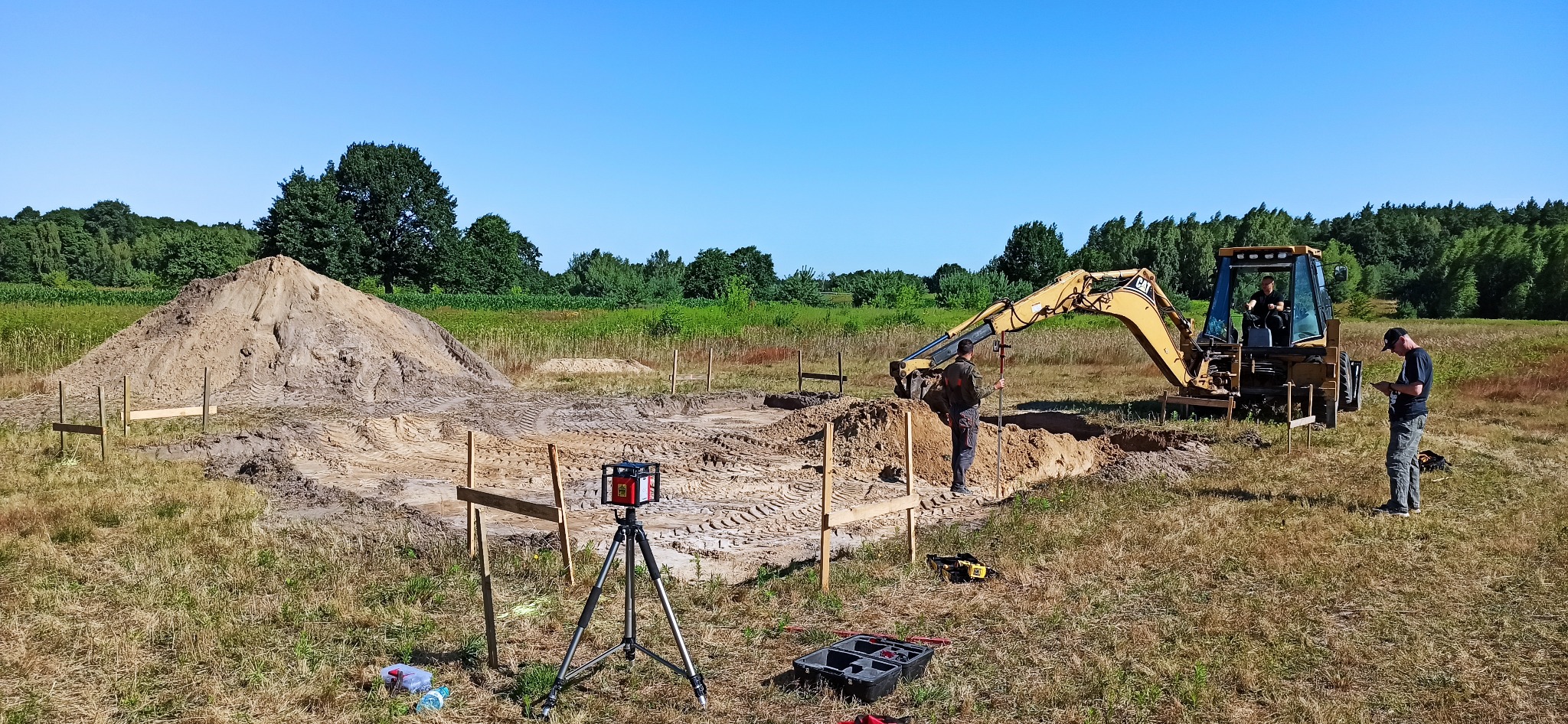 Przygotowanie terenu pod budowę: wykop fundamentów z użyciem koparko-ładowarki, wyznaczanie poziomu za pomocą niwelatora laserowego na statywie, widoczny stos ziemi, drewniane szalunki.