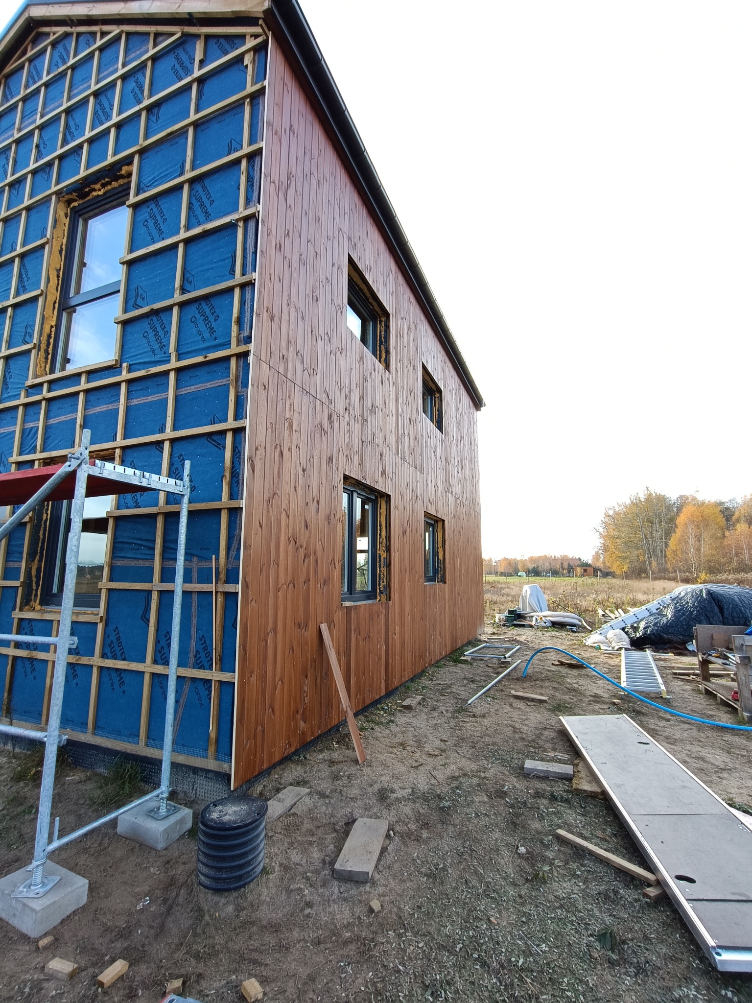 Dwuetapowa budowa domu szkieletowego: ocieplenie z membraną i drewniana elewacja. Widoczne rusztowanie i teren budowy. Ujęcie z dołu, perspektywa zbieżna.
