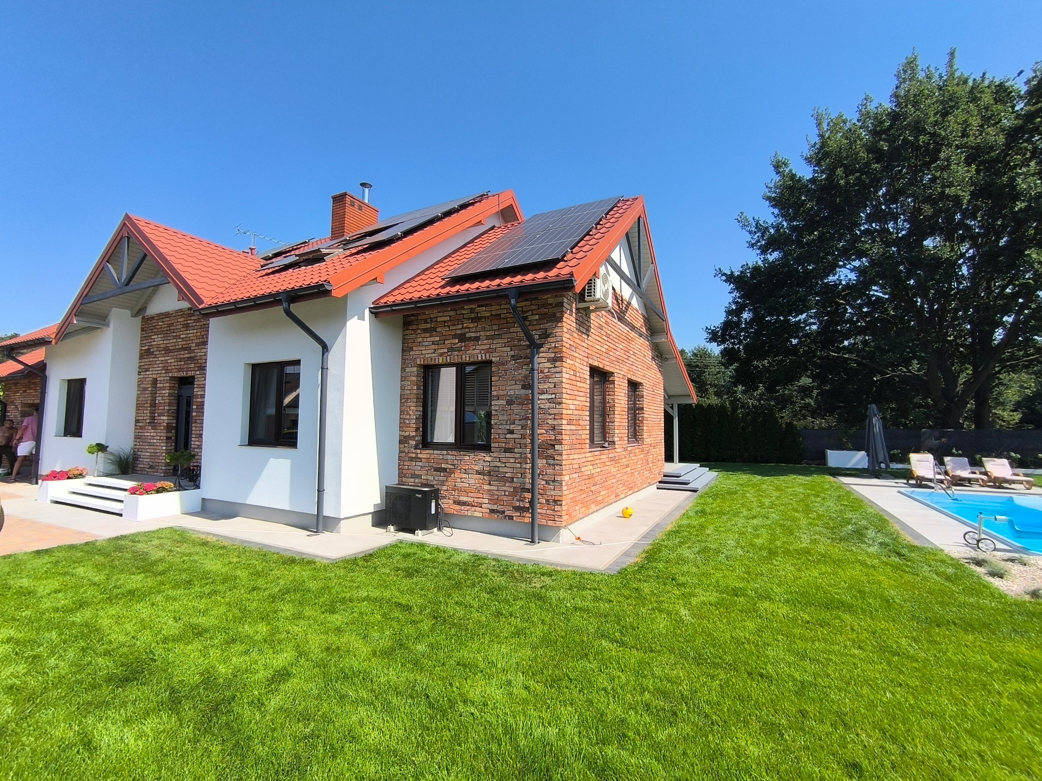 Nowoczesny dom z ceglaną elewacją, panelami słonecznymi na czerwonym dachu i zadbanym trawnikiem. W tle basen i drzewa. Jasne, słoneczne ujęcie.