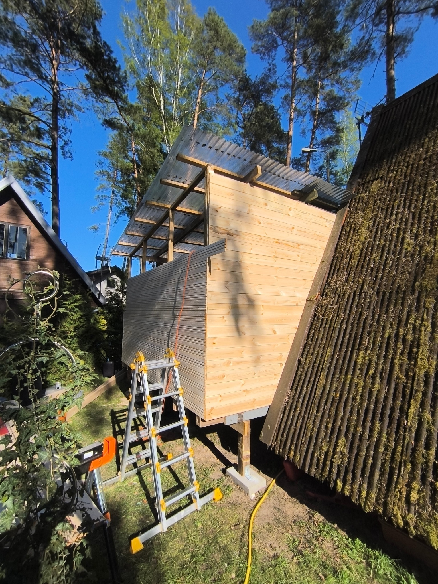 Wznoszenie drewnianej konstrukcji z przezroczystym zadaszeniem na tle drzew i błękitnego nieba. Widoczna drabina i narzędzia. Częściowo ukończona budowa.
