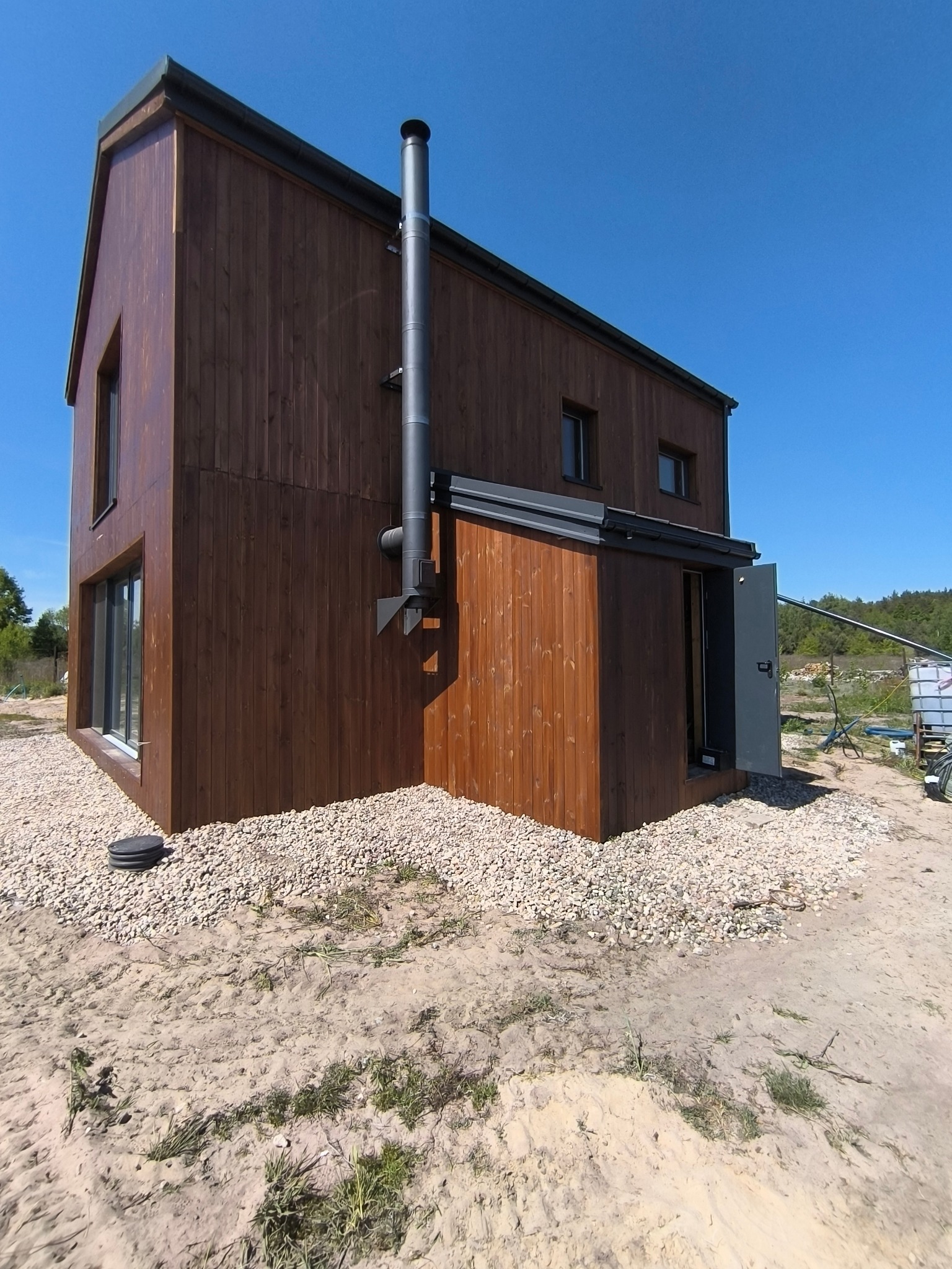 Nowoczesny dom szkieletowy z ciemnego drewna, z kominem i otwartymi drzwiami, otoczony żwirem i piaskiem w słoneczny dzień. Widok z dołu.