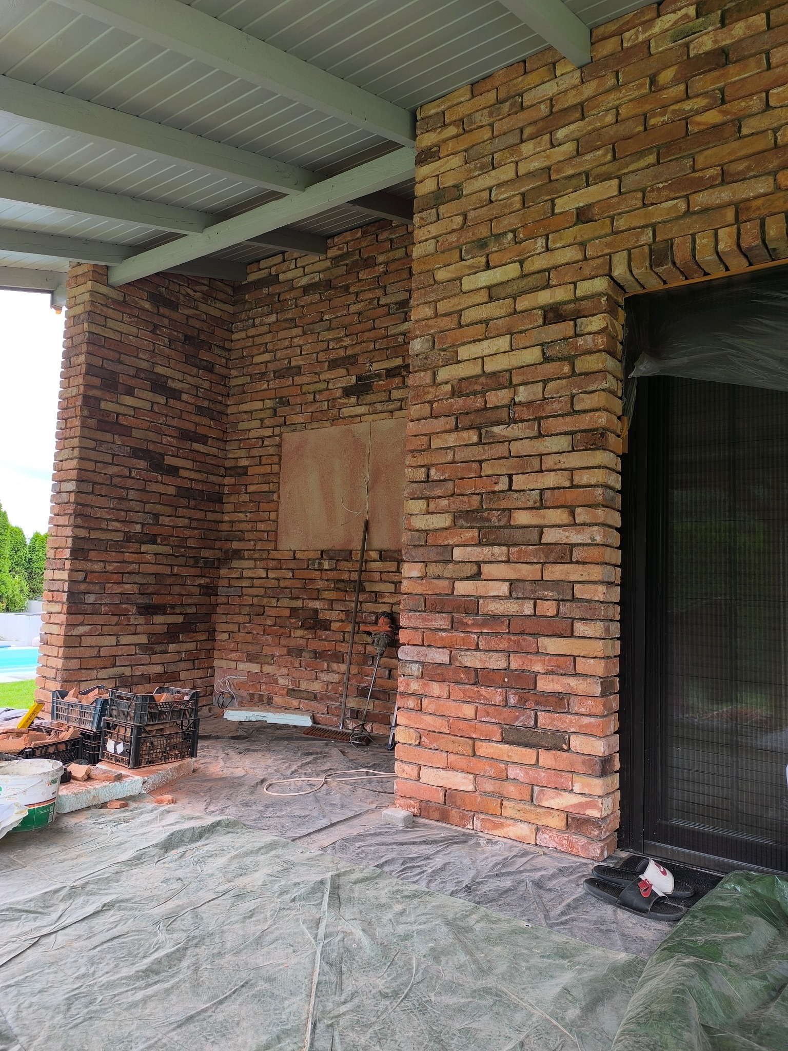 Murowana ściana z cegły klinkierowej pod zadaszeniem, widoczne narzędzia i materiały budowlane, folia ochronna na podłodze. Etap prac wykończeniowych.