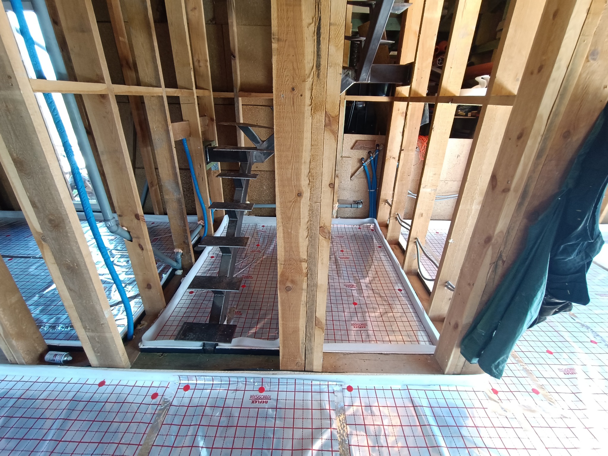 Konstrukcja drewniana poddasza z instalacją ogrzewania podłogowego na folii refleksyjnej z czerwoną siatką, widoczne rury instalacyjne i fragment schodów.