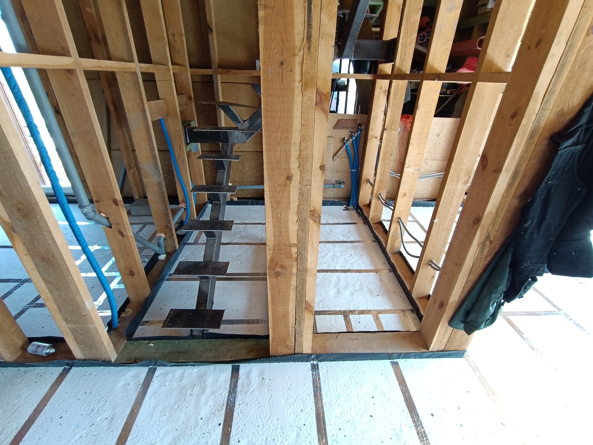 Wnętrze domu w trakcie budowy szkieletowej: drewniana konstrukcja, izolacja podłogi, instalacje wodne. Widoczne schody metalowe i elementy konstrukcyjne dachu.